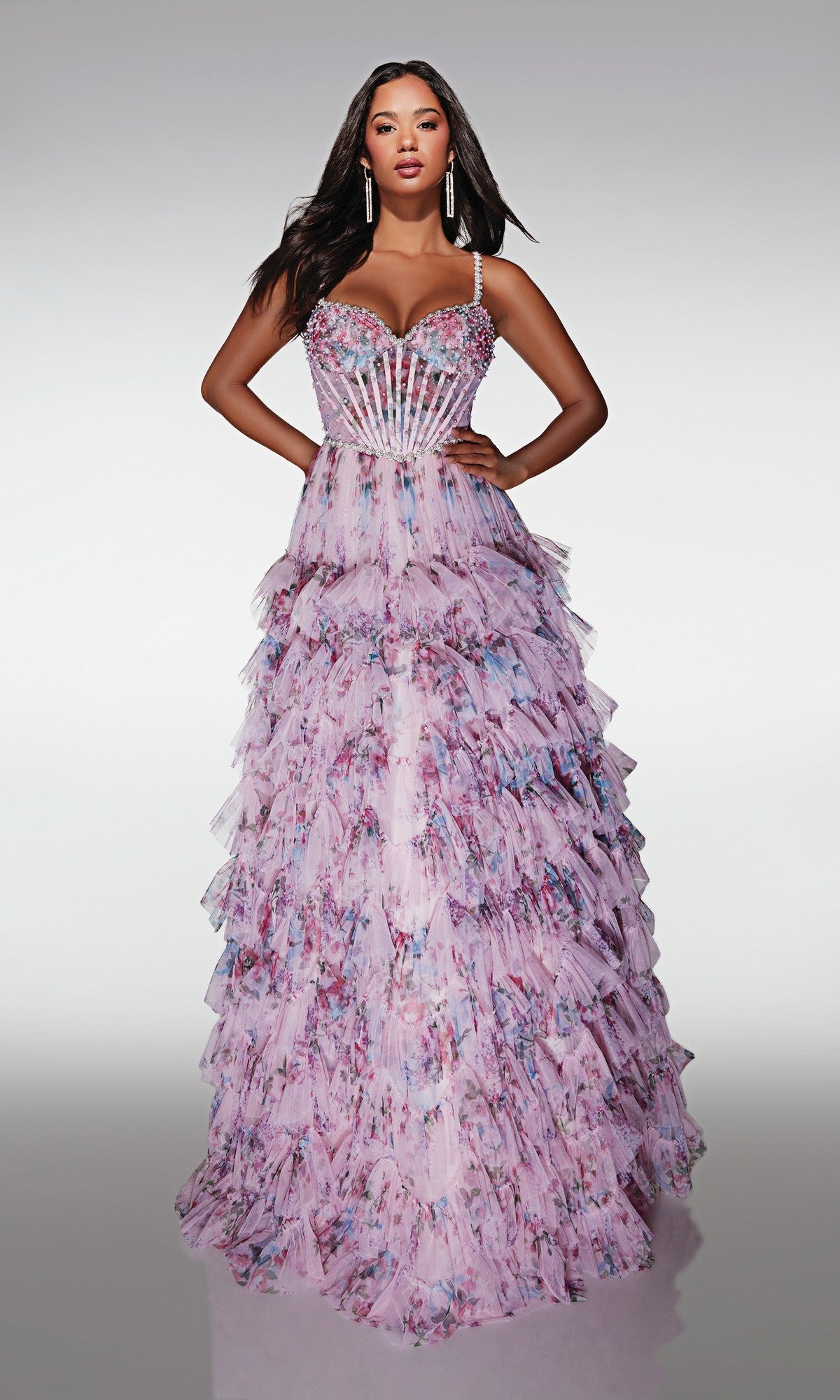 A woman poses in the Alyce 62182 long prom dress, featuring a tiered floral ball gown skirt, embellished straps, and a fitted sheer corset bodice, set against a plain light background.